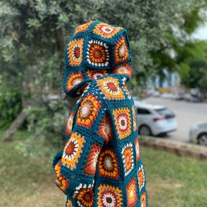 K&ouml;nnte beinhalten: Eine tealblaue geh&auml;kelte Kapuzenjacke mit einem geometrischen Muster aus orangefarbenen, braunen und wei&szlig;en Quadraten.