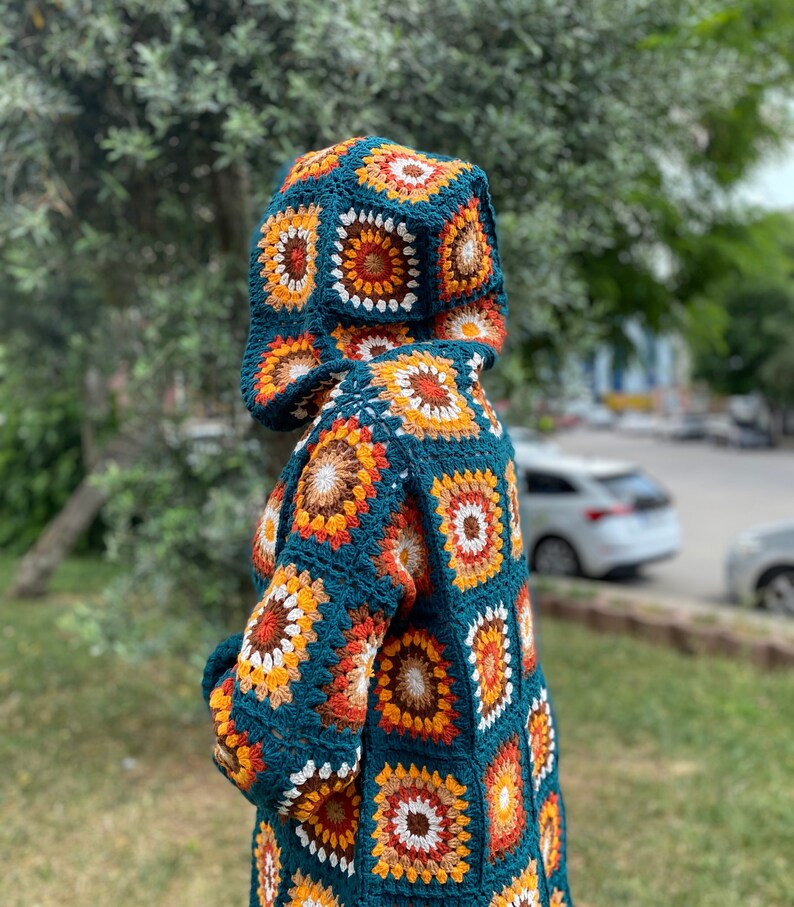 K&ouml;nnte beinhalten: Eine tealblaue geh&auml;kelte Kapuzenjacke mit einem geometrischen Muster aus orangefarbenen, braunen und wei&szlig;en Quadraten.