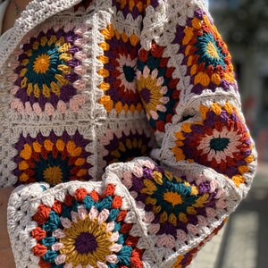 Puede incluir: Un c&aacute;rdigan tejido a mano con un patr&oacute;n de cuadrados de la abuela. La prenda muestra una vibrante combinaci&oacute;n de colores turquesa, naranja, amarillo, morado y rosa, sobre un fondo beige neutro. Las mangas y el cuerpo est&aacute;n hechos de cuadrados individuales.