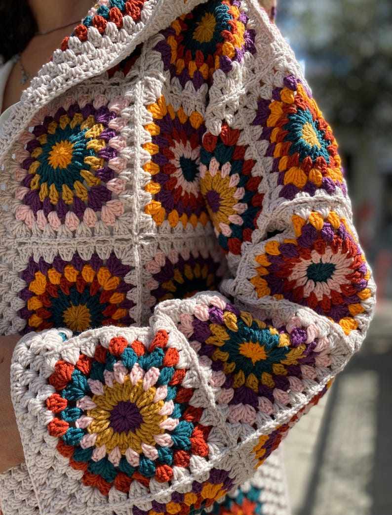 Puede incluir: Un c&aacute;rdigan tejido a mano con un patr&oacute;n de cuadrados de la abuela. La prenda muestra una vibrante combinaci&oacute;n de colores turquesa, naranja, amarillo, morado y rosa, sobre un fondo beige neutro. Las mangas y el cuerpo est&aacute;n hechos de cuadrados individuales.