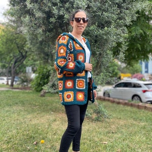 K&ouml;nnte beinhalten: Ein tealblauer H&auml;kelcardigan mit Kapuze, der ein farbenfrohes geometrisches Muster aus orangefarbenen, gelben und wei&szlig;en Quadraten aufweist. Der Cardigan wird von einer Person mit schwarzer Hose und Sonnenbrille getragen.