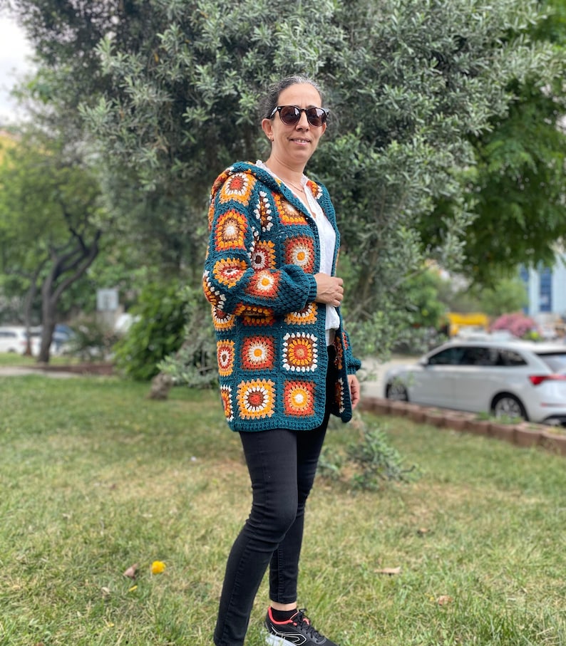 K&ouml;nnte beinhalten: Ein tealblauer H&auml;kelcardigan mit Kapuze, der ein farbenfrohes geometrisches Muster aus orangefarbenen, gelben und wei&szlig;en Quadraten aufweist. Der Cardigan wird von einer Person mit schwarzer Hose und Sonnenbrille getragen.
