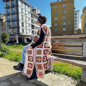 May include: A woman wearing a crocheted vest with a colorful geometric pattern sits on a wooden bench. The vest is made of white, pink, orange, yellow, green, and brown yarn. The woman is wearing blue jeans and black boots.