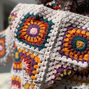 Puede incluir: Primer plano de un su&eacute;ter con capucha de ganchillo. El su&eacute;ter presenta un dise&ntilde;o de cuadrados de la abuela con una paleta de colores beige, morado, naranja, verde azulado y amarillo. La capucha est&aacute; levantada y el su&eacute;ter parece hecho a mano.