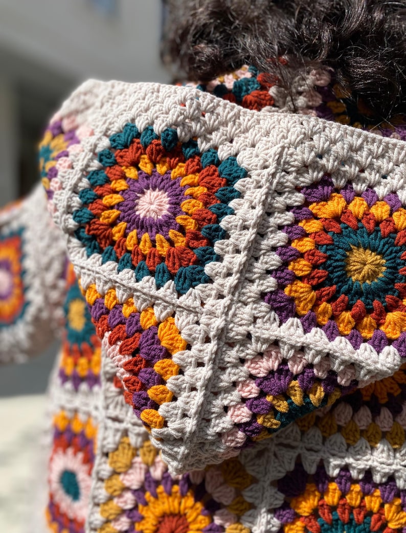 Puede incluir: Primer plano de un su&eacute;ter con capucha de ganchillo. El su&eacute;ter presenta un dise&ntilde;o de cuadrados de la abuela con una paleta de colores beige, morado, naranja, verde azulado y amarillo. La capucha est&aacute; levantada y el su&eacute;ter parece hecho a mano.