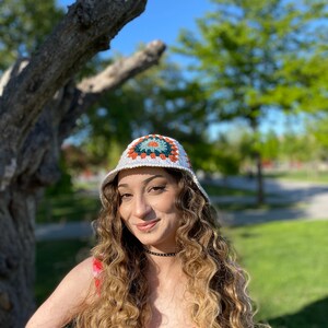 Puede incluir: Una mujer con el pelo largo y rizado lleva un sombrero de cubo de crochet blanco con un patr&oacute;n geom&eacute;trico colorido. Lleva un vestido de estampado floral rosa y blanco.