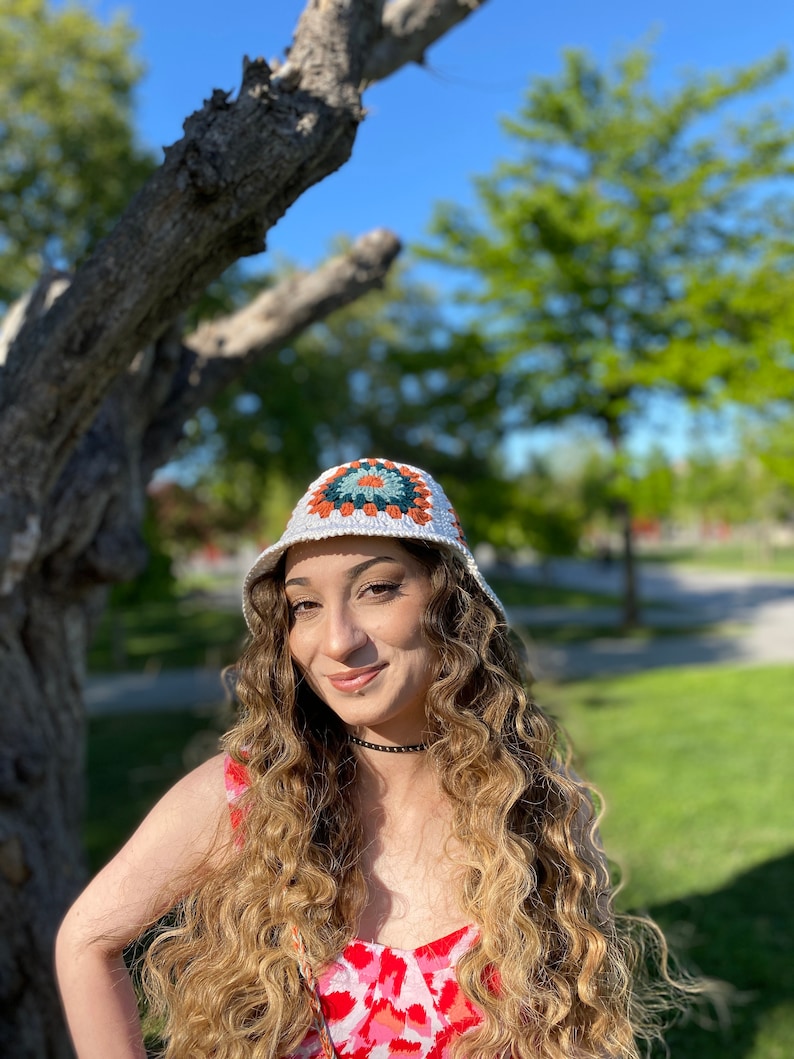 Puede incluir: Una mujer con el pelo largo y rizado lleva un sombrero de cubo de crochet blanco con un patr&oacute;n geom&eacute;trico colorido. Lleva un vestido de estampado floral rosa y blanco.