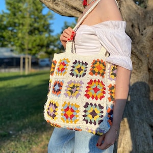 May include: A white crochet tote bag with a colorful geometric pattern. The bag has a long strap and is made of thick yarn.