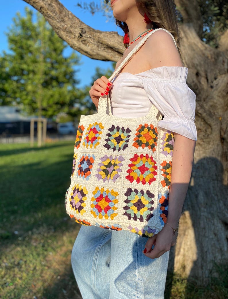 May include: A white crochet tote bag with a colorful geometric pattern. The bag has a long strap and is made of thick yarn.