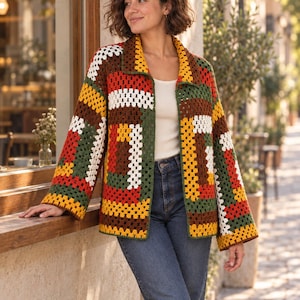 May include: A long-sleeved crochet cardigan featuring a multi-coloured granny square pattern in shades of brown, dark green, mustard yellow, red, and white. The garment has a collared neckline, an open front, and a loose, boxy fit with wide sleeves.