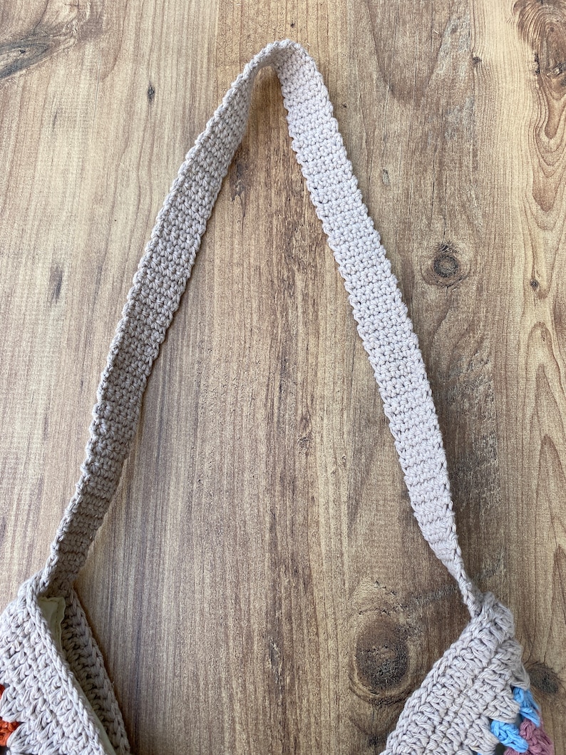 May include: A beige crochet tote bag with a long strap, set against a wooden backdrop. The bag showcases a granny square pattern with visible stitching and a robust strap. The neutral colour scheme of the bag makes it adaptable for various uses.