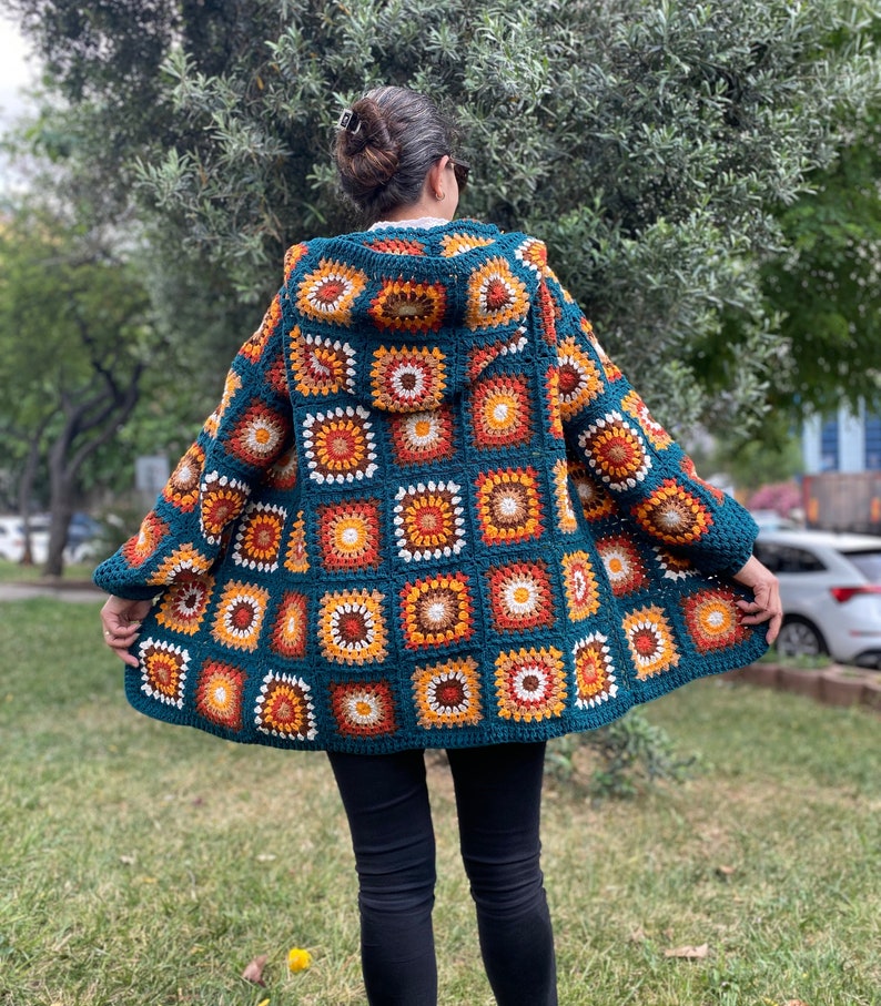 K&ouml;nnte beinhalten: Eine tealblaue geh&auml;kelte Strickjacke mit Kapuze, die ein farbenfrohes geometrisches Muster aus orangefarbenen, gelben, wei&szlig;en und braunen Quadraten aufweist. Die Jacke hat eine lockere Passform und lange &Auml;rmel.