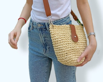 May include: A light brown woven crossbody bag with a brown leather strap and a brown leather flap with a button closure. The bag is worn over the shoulder of a person wearing blue jeans and a white t-shirt.
