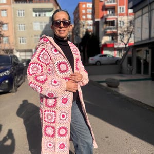 Könnte beinhalten: Langer, kapuzenbesetzter Häkelcardigan in Rosa-, Weiß- und Rosatönen. Das offene Design hat ein geometrisches Muster aus Quadraten und Kreisen. Der Cardigan wird an einem sonnigen Tag im Freien getragen.
