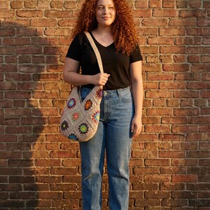 May include: A beige crochet tote bag with colourful floral squares. The bag has a long strap. The person is wearing a black V-neck shirt and blue jeans. The background is a brick wall.