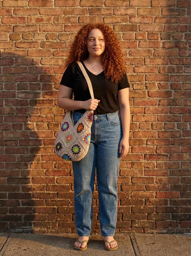 May include: A beige crochet tote bag with colourful floral squares. The bag has a long strap. The person is wearing a black V-neck shirt and blue jeans. The background is a brick wall.