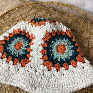 Puede incluir: Un sombrero de cubo de crochet blanco con un patr&oacute;n geom&eacute;trico colorido en tonos naranja, verde y azul.