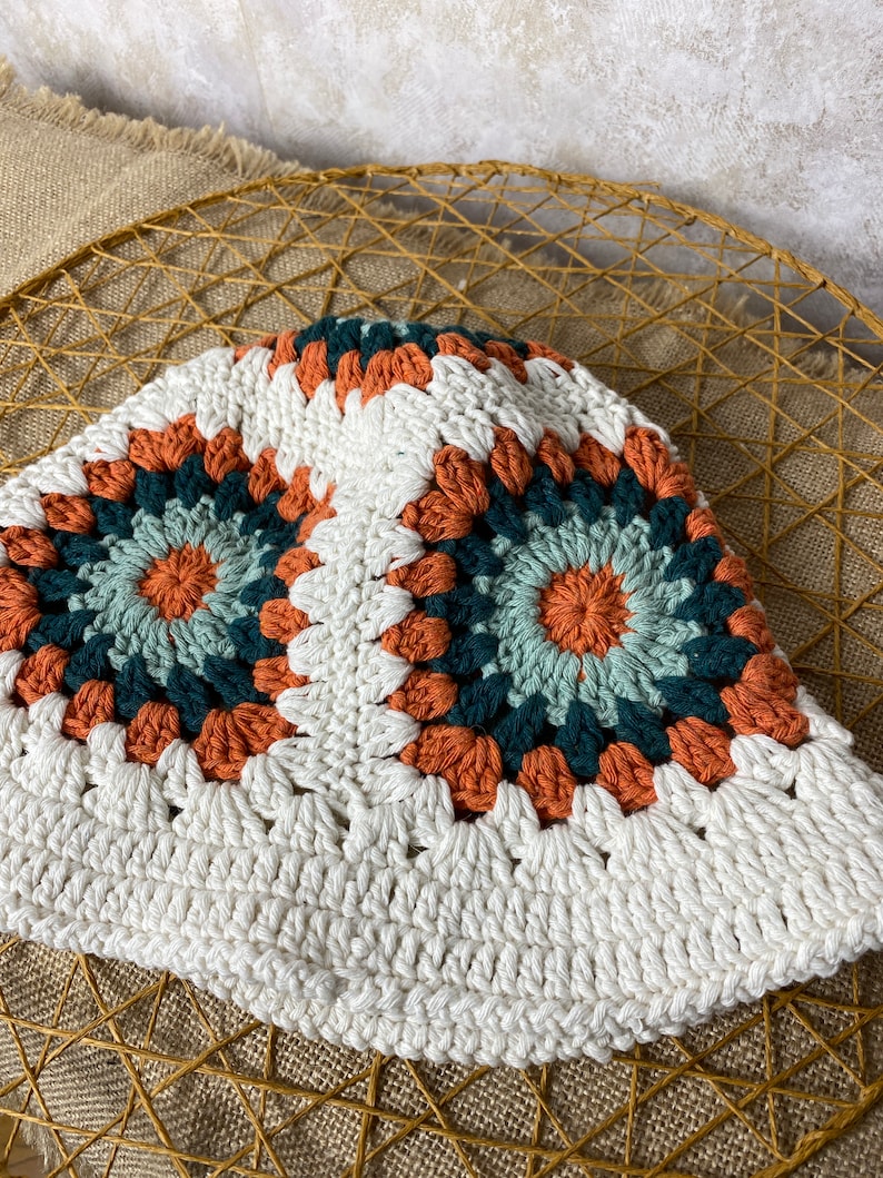 Puede incluir: Un sombrero de cubo de crochet blanco con un patr&oacute;n geom&eacute;trico colorido en tonos naranja, verde y azul.