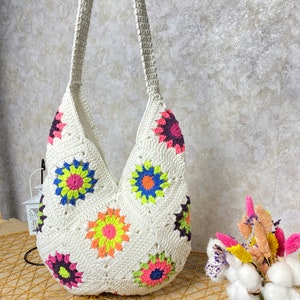 Op de afbeelding: Witte gehaakte tote bag met een kleurrijk bloemenpatroon. De tas heeft een lange band en is gemaakt van katoenen garen.