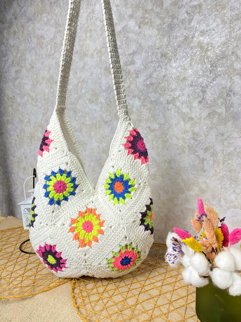 Op de afbeelding: Witte gehaakte tote bag met een kleurrijk bloemenpatroon. De tas heeft een lange band en is gemaakt van katoenen garen.