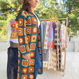 May include: A woman wearing a long, green crochet cardigan with a colorful geometric pattern. The cardigan is made of granny squares and has a relaxed fit. The woman is standing in front of a rack of similar cardigans in various colors.