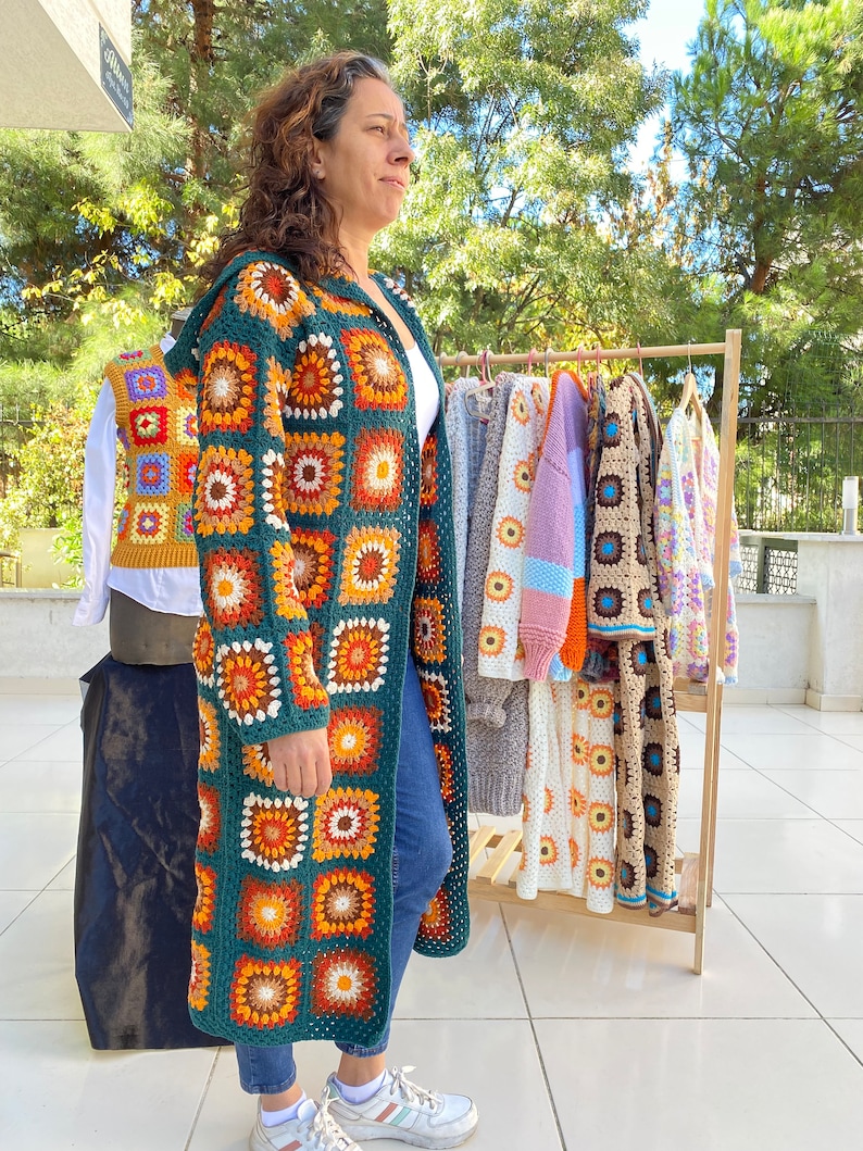 May include: A woman wearing a long, green crochet cardigan with a colorful geometric pattern. The cardigan is made of granny squares and has a relaxed fit. The woman is standing in front of a rack of similar cardigans in various colors.