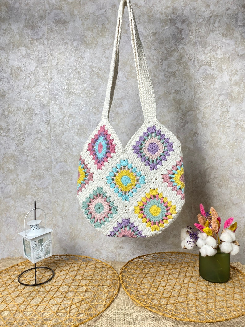 May include: A white crocheted tote bag with a round bottom and colorful floral designs. The bag has two long straps and is hanging from a hook.