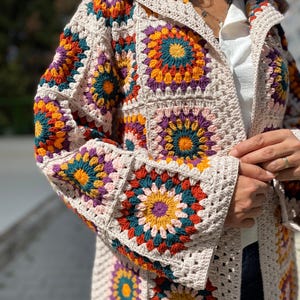 Puede incluir: Una chaqueta de crochet con capucha de color crema con patrones cuadrados coloridos. Los cuadrados presentan dise&ntilde;os florales vibrantes en tonos naranja, amarillo, verde azulado y morado. La chaqueta tiene mangas largas y un corte holgado.