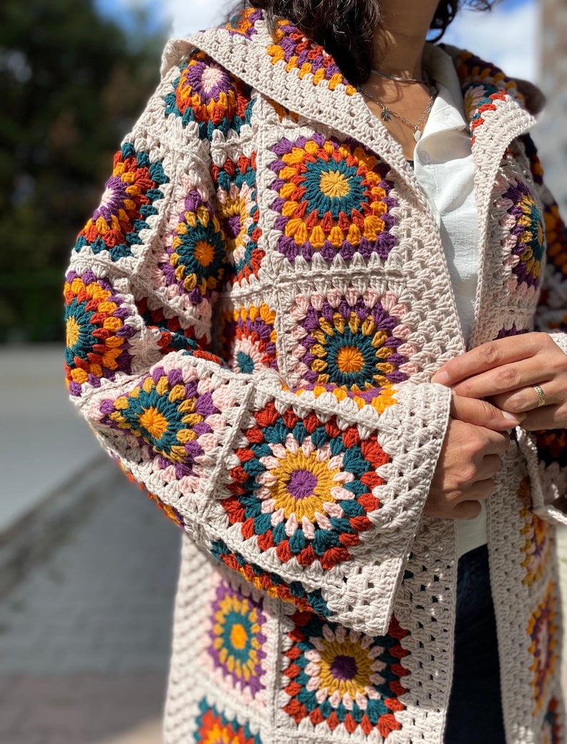 Puede incluir: Una chaqueta de crochet con capucha de color crema con patrones cuadrados coloridos. Los cuadrados presentan dise&ntilde;os florales vibrantes en tonos naranja, amarillo, verde azulado y morado. La chaqueta tiene mangas largas y un corte holgado.