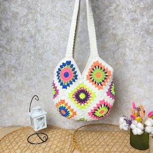 May include: White crochet tote bag with colorful floral designs. The bag has a long strap and is made of a soft, lightweight material.