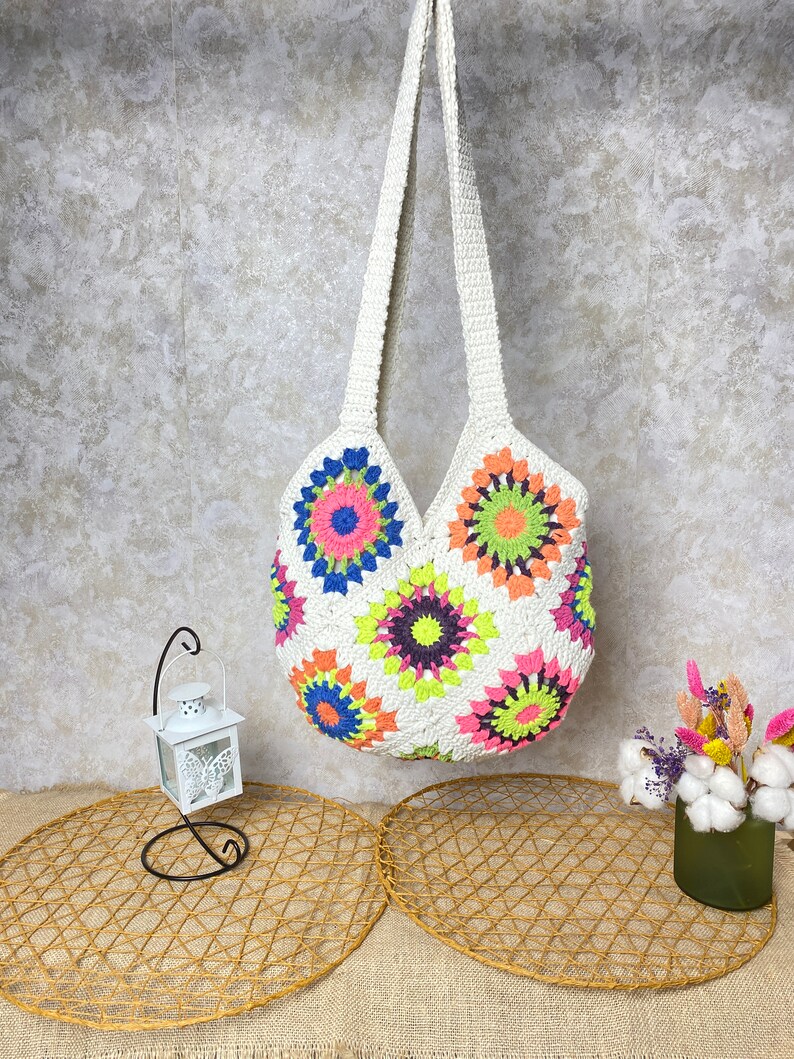 May include: White crochet tote bag with colorful floral designs. The bag has a long strap and is made of a soft, lightweight material.