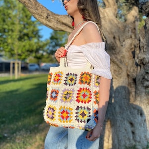 May include: A white crochet tote bag with colorful square patterns. The bag has a wide, flat base and two handles. The bag is made of a soft, textured yarn.
