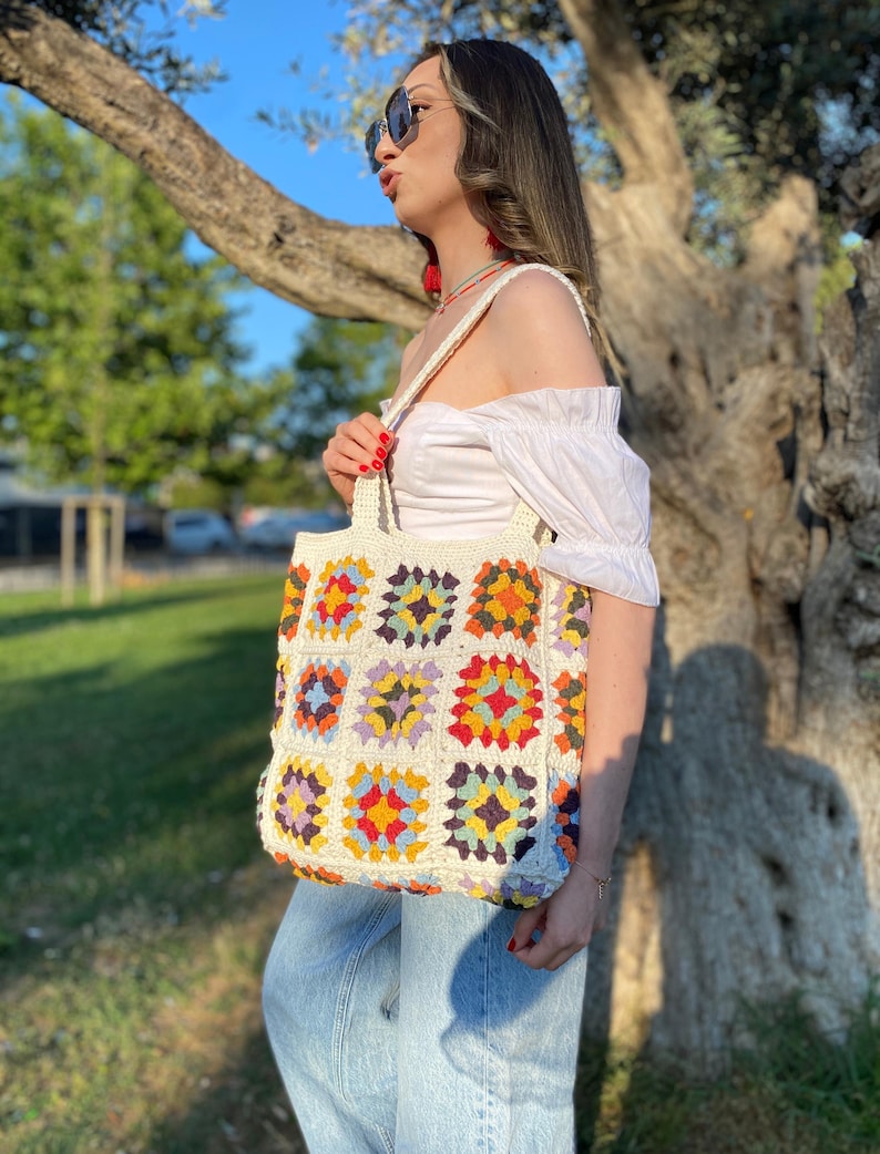 May include: A white crochet tote bag with colorful square patterns. The bag has a wide, flat base and two handles. The bag is made of a soft, textured yarn.