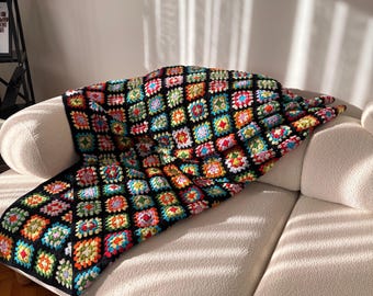 May include: A colorful, handmade crochet blanket draped over a cream-colored sofa. The blanket features a black background with vibrant squares of red, yellow, blue, green, and orange. The blanket's texture and pattern are clearly visible, suggesting a cozy, handcrafted item.
