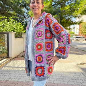 May include: A gray crocheted cardigan with a hood, featuring a colorful geometric pattern of squares with pink, orange, green, and purple accents.