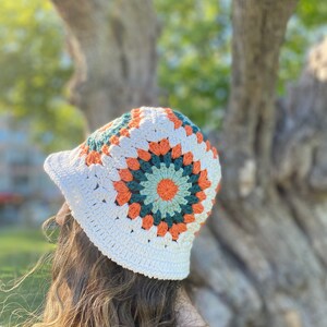 Puede incluir: Un sombrero de cubo de crochet blanco con un dise&ntilde;o de cuadrado de la abuela colorido en tonos naranja, verde y blanco.