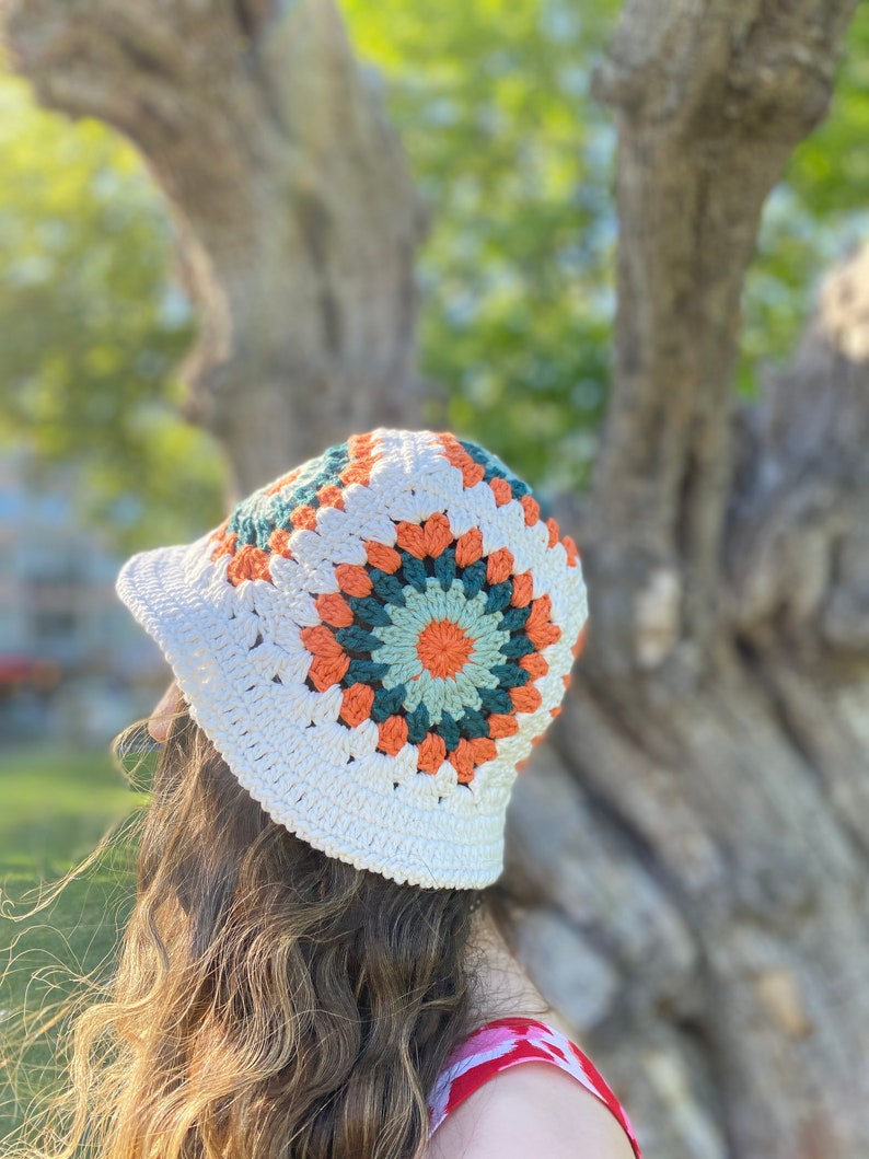 Puede incluir: Un sombrero de cubo de crochet blanco con un dise&ntilde;o de cuadrado de la abuela colorido en tonos naranja, verde y blanco.