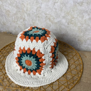 Puede incluir: Un sombrero de cubo blanco tejido a crochet con un patr&oacute;n geom&eacute;trico colorido en tonos naranja, verde y azul.