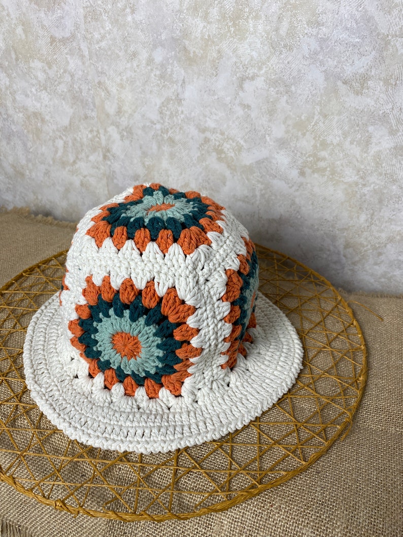 Puede incluir: Un sombrero de cubo blanco tejido a crochet con un patr&oacute;n geom&eacute;trico colorido en tonos naranja, verde y azul.