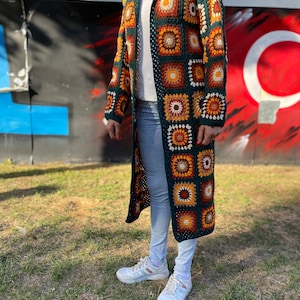 May include: A long, green crochet cardigan with a hood. The cardigan features a colorful geometric pattern with orange, brown, and white accents. The cardigan is worn over a white shirt and blue jeans.