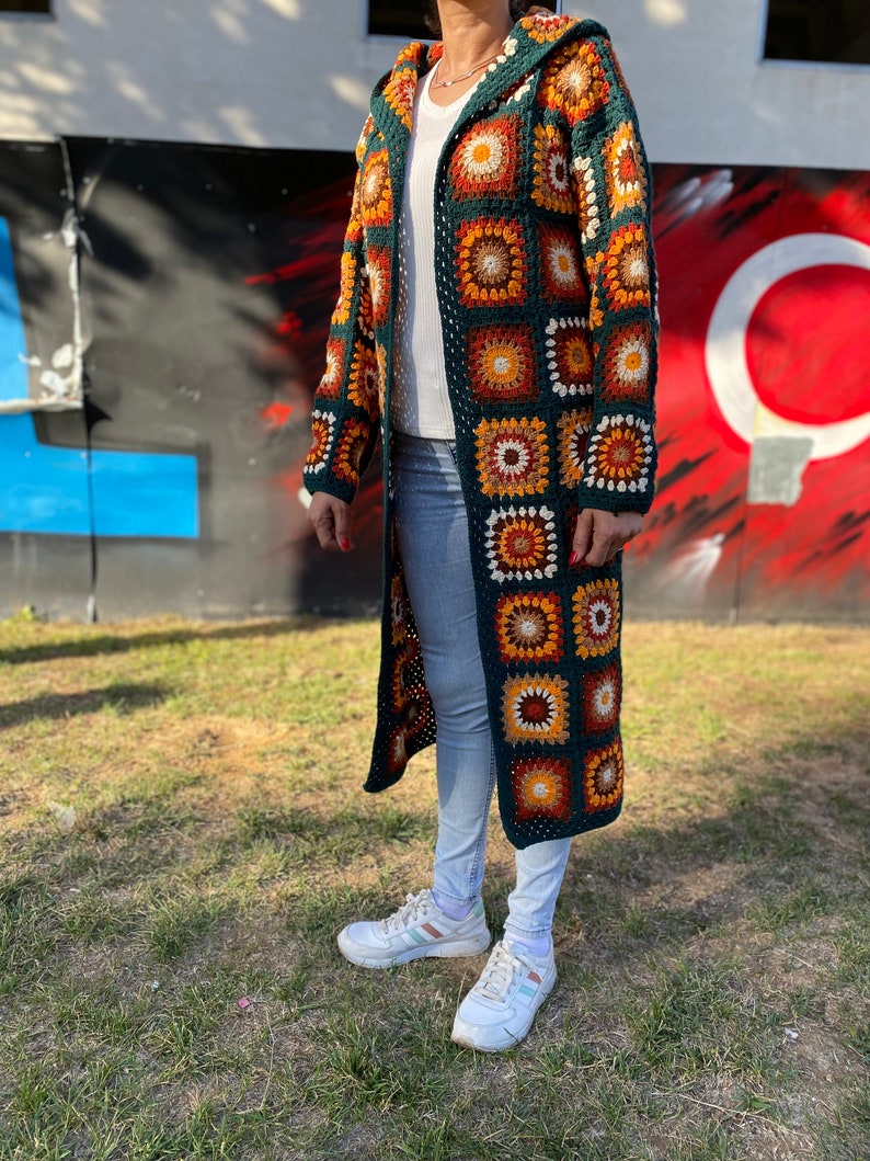 May include: A long, green crochet cardigan with a hood. The cardigan features a colorful geometric pattern with orange, brown, and white accents. The cardigan is worn over a white shirt and blue jeans.