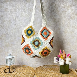 May include: A white crocheted tote bag with a colorful geometric pattern. The bag has a single long strap and is hanging from a wall.