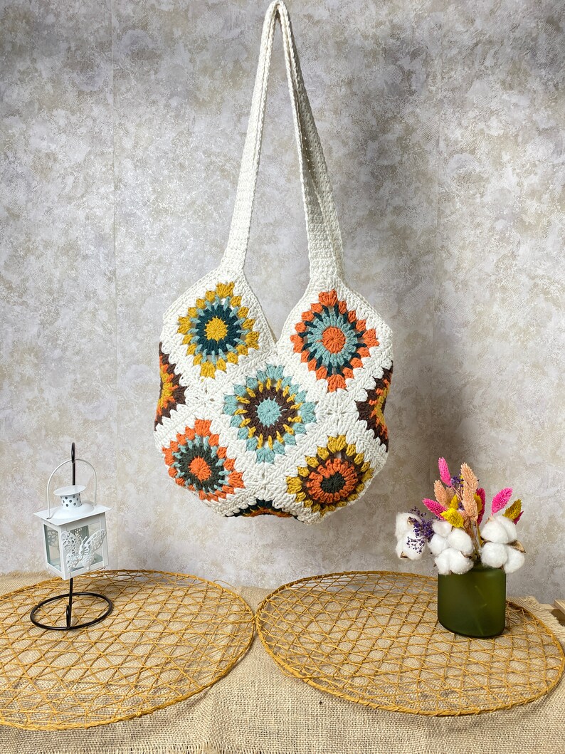 May include: A white crocheted tote bag with a colorful geometric pattern. The bag has a single long strap and is hanging from a wall.