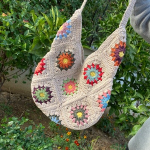 May include: A beige crochet shoulder bag with colourful floral designs. The bag features a patchwork design with squares of beige yarn, each adorned with a different flower in shades of red, orange, yellow, blue, and green. The bag has a long strap.