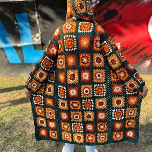 May include: A long, green crocheted cardigan with a hood. The cardigan is made of squares with a colorful pattern of orange, brown, and white.