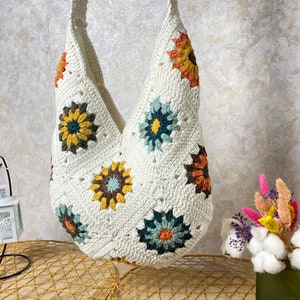 Op de afbeelding: Witte gehaakte tote bag met kleurrijke bloemenmotieven. De tas heeft een brede, open bovenkant en een lange band.