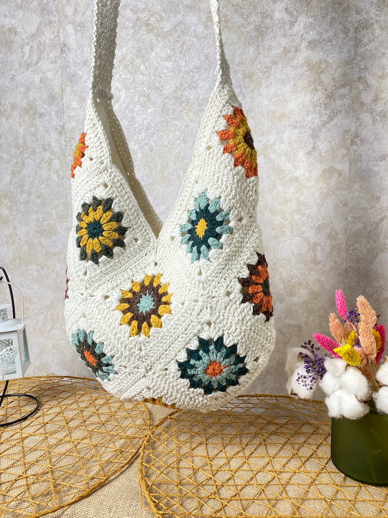Op de afbeelding: Witte gehaakte tote bag met kleurrijke bloemenmotieven. De tas heeft een brede, open bovenkant en een lange band.
