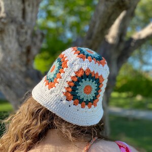 Puede incluir: Sombrero de cubo de crochet blanco con un patr&oacute;n geom&eacute;trico colorido en tonos naranja, verde y blanco.