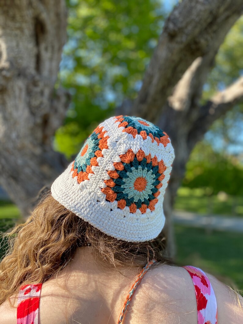 Puede incluir: Sombrero de cubo de crochet blanco con un patr&oacute;n geom&eacute;trico colorido en tonos naranja, verde y blanco.