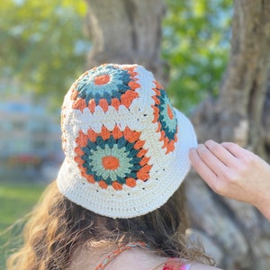 Puede incluir: Un sombrero de cubo de crochet blanco con un patr&oacute;n geom&eacute;trico colorido en tonos naranja, verde y blanco.